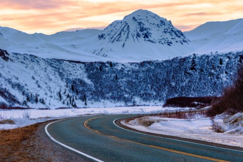 A winding road leads through a snowy landscape, with a majestic mountain in the background under a colorful sunset.