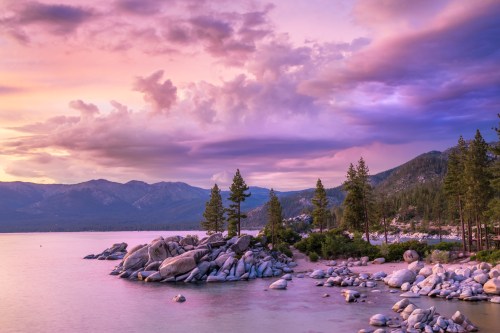 A serene lakeside scene at sunset, featuring rocky shores, pine trees, and colorful clouds over the mountains.