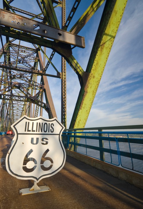 A weathered US Route 66 sign stands on a bridge, with green metal supports and a blue sky in the background.