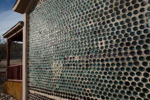A wall covered in colorful glass bottles, creating a mosaic effect, with a wooden structure in the foreground.