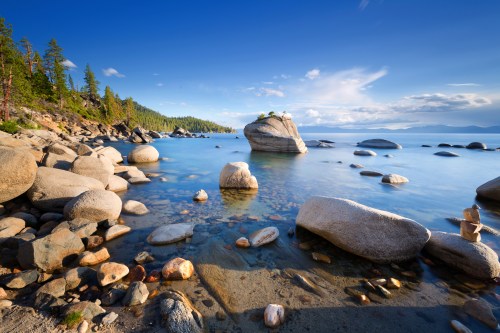 A serene lakeside scene with smooth rocks, clear water, and a blue sky dotted with clouds, surrounded by trees.