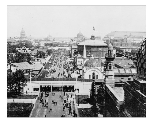 A historic black-and-white view of a bustling fairground with buildings and crowds, showcasing early 20th-century architecture.