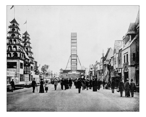 Historic street scene with crowds, buildings, and a large Ferris wheel in the background. Black and white photo.