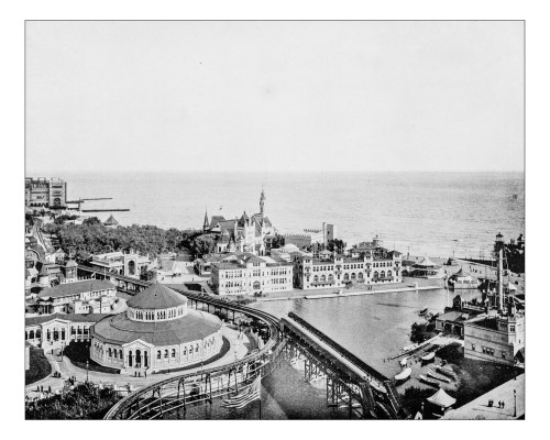 Aerial view of a coastal amusement park with rides, buildings, and a body of water in the background.