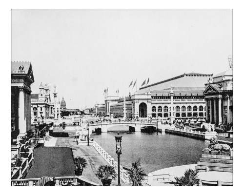 Historic black-and-white view of a grand canal with buildings and flags, showcasing architectural details and a bridge.