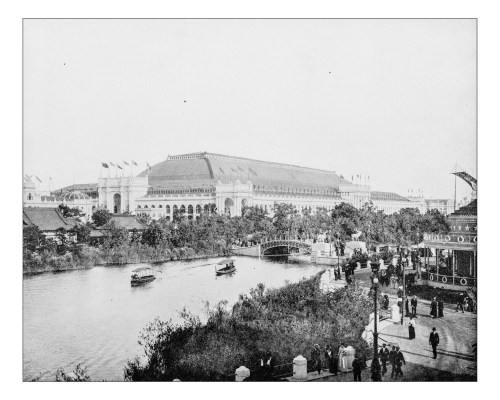 Historic black-and-white image of a large building by a river, with boats and people in a park-like setting.