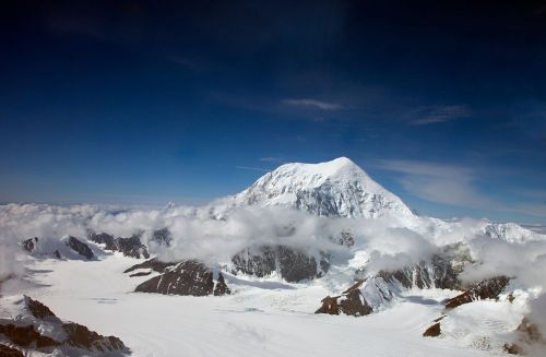 A snow-capped mountain rises above clouds, surrounded by rugged terrain and a clear blue sky.