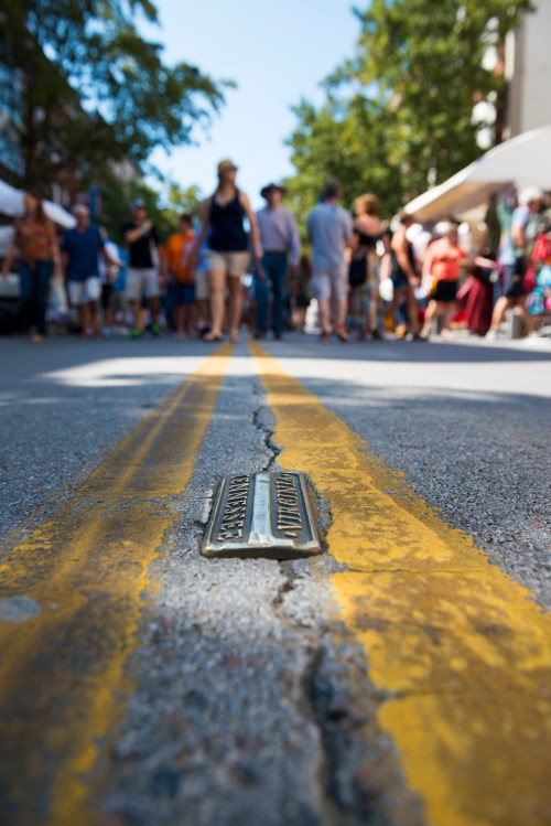 A close-up of a cracked street with a metal plaque, blurred crowds and tents in the background during a street event.
