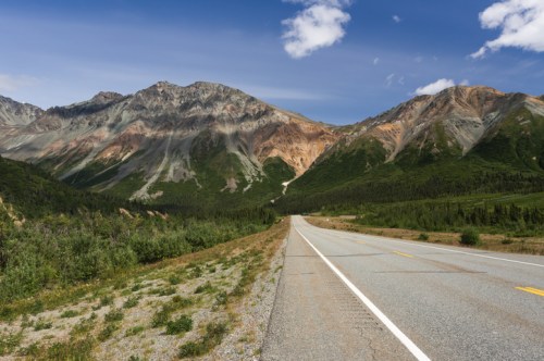 A winding road leads through a lush green valley, flanked by colorful mountains under a clear blue sky.