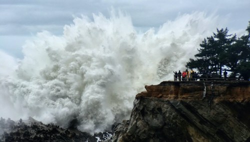 A massive wave crashes against a rocky shore, with people observing from a nearby viewpoint.