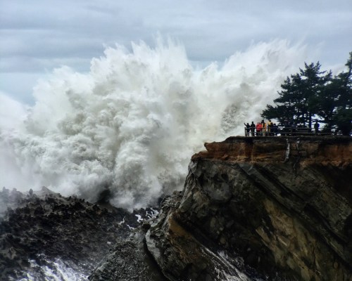 A large wave crashes against a rocky cliff, with people standing safely at the edge, watching the powerful surf.