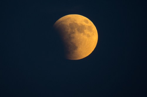 A partially eclipsed moon glowing in a dark sky, with a warm orange hue on one side.