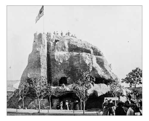 Historic black and white photo of a rock formation with people on top, surrounded by trees and a flag.
