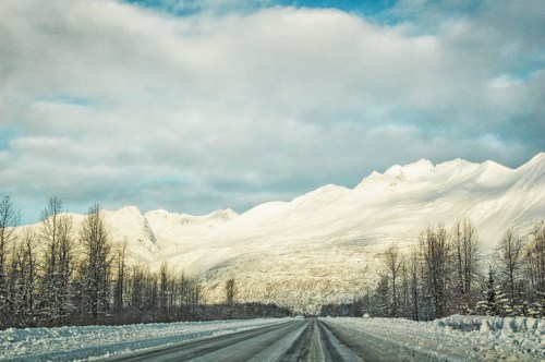 A snowy road leads towards majestic mountains under a cloudy sky, surrounded by bare trees.