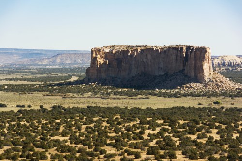 A large, flat-topped rock formation rises above a vast, green landscape under a clear blue sky.