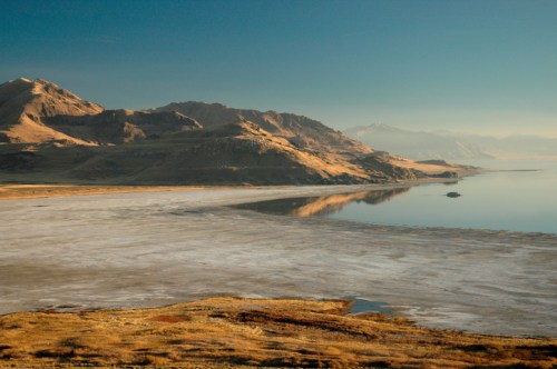 A serene landscape featuring mountains, a calm lake, and a salt flat under a clear blue sky.
