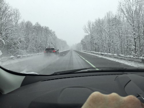A snowy road scene viewed from a car, with trees covered in snow and another vehicle in the distance.