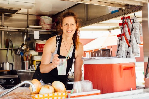 A smiling woman in an apron holds a drink at a food truck, surrounded by cooking equipment and supplies.