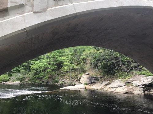 A view under a stone bridge, with a calm river and lush green trees along the rocky banks.