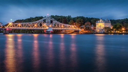 A illuminated bridge spans a calm river at dusk, with trees and a historic building in the background.