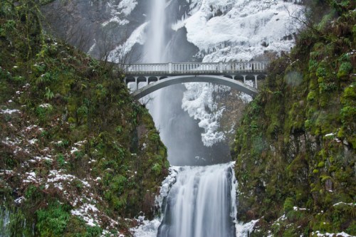 A snowy waterfall cascades beneath a stone bridge, surrounded by lush greenery and rocky cliffs.