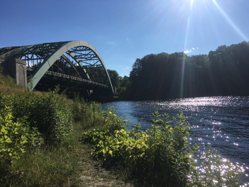 A bridge arches over a shimmering river, surrounded by lush greenery and a clear blue sky.
