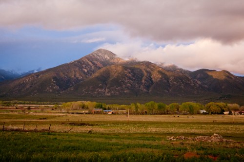 A scenic view of a mountain range under cloudy skies, with green fields and trees in the foreground.