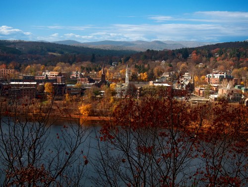 A scenic view of a town by a river, surrounded by autumn foliage and mountains under a clear blue sky.