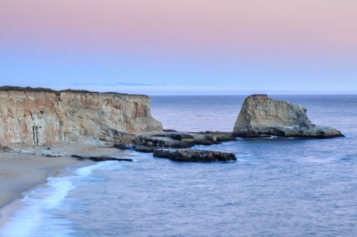 A serene coastal scene featuring cliffs, rocky formations, and gentle waves under a pastel sky at sunset.