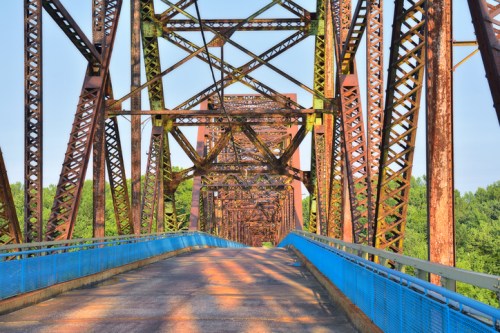 Rusty metal bridge with intricate beams and a blue railing, surrounded by greenery under a clear sky.