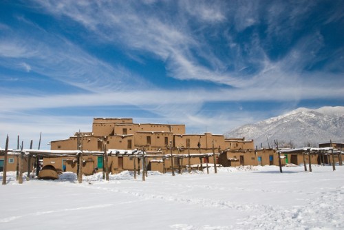 A snow-covered landscape featuring adobe buildings under a blue sky with wispy clouds and mountains in the background.