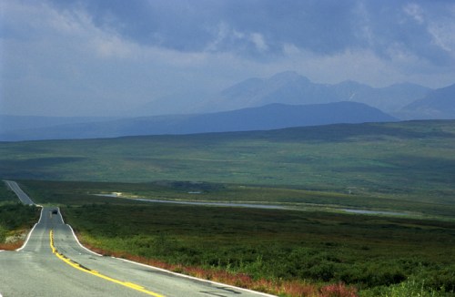 A winding road stretches through a vast green landscape, with mountains in the distance under a cloudy sky.