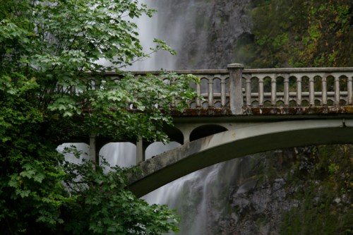 A stone bridge with a decorative railing spans a lush, green landscape near a cascading waterfall.
