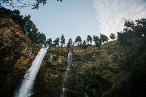 A waterfall cascades down a rocky cliff, surrounded by trees and a clear blue sky.