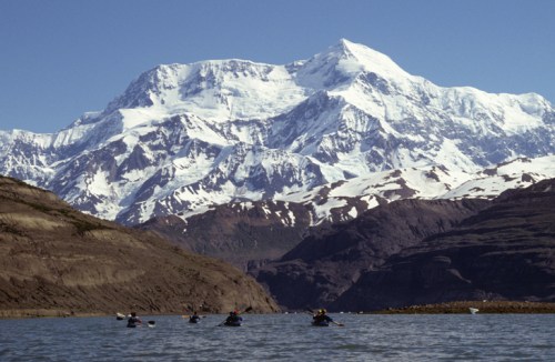 A group of kayakers paddles on a calm lake with snow-capped mountains in the background under a clear blue sky.