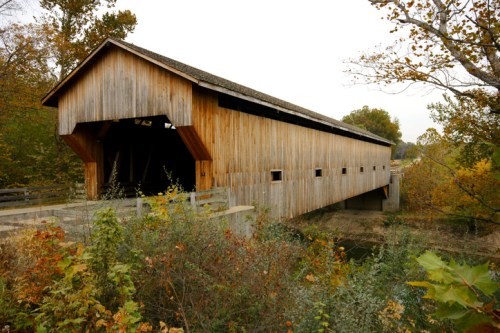 A wooden covered bridge surrounded by trees and foliage, set against a cloudy sky.