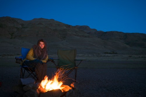 A person sits by a campfire in the evening, surrounded by mountains and two empty camping chairs.