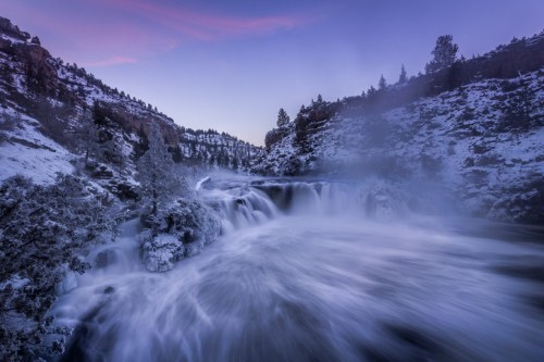 A serene waterfall cascades through a snowy landscape at dusk, surrounded by trees and mist.