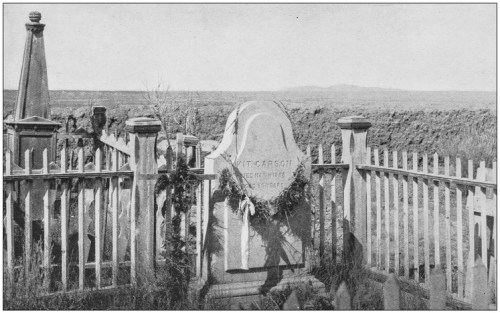 Gravestone of Kit Carson surrounded by a wooden fence, with a desert landscape in the background.