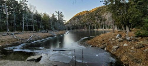 A serene lake surrounded by trees and mountains, with patches of ice on the water and a clear sky above.