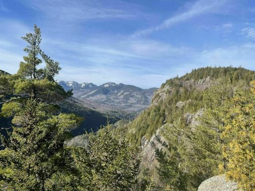 A scenic view of mountains and trees under a clear blue sky, showcasing rugged terrain and distant peaks.