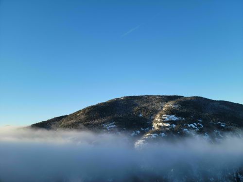 A mountain peak rises above a blanket of fog under a clear blue sky. Snow is visible on the slopes.