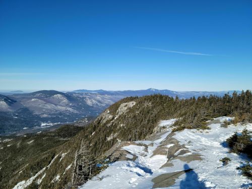 A panoramic view of snow-covered mountains under a clear blue sky, with rocky terrain in the foreground.