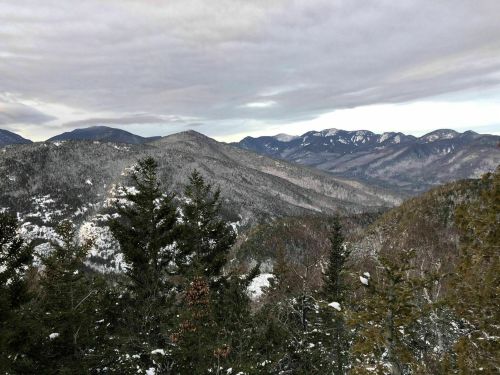 A snowy mountain landscape with evergreen trees in the foreground and distant peaks under a cloudy sky.