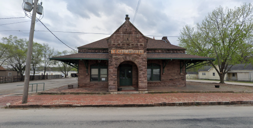 Historic stone train station with a peaked roof, surrounded by trees and a gravel lot under a cloudy sky.