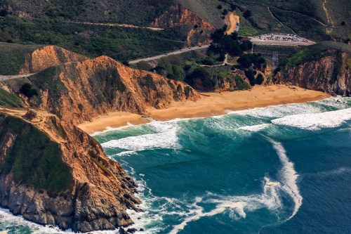 Aerial view of a sandy beach surrounded by cliffs and waves, with a road and parking area in the background.