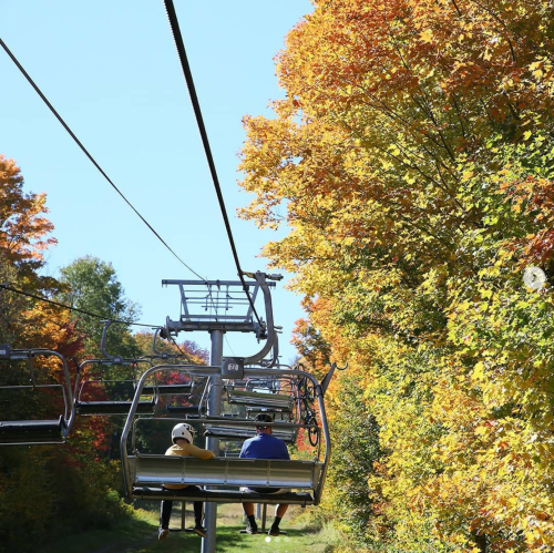 Two people ride a chairlift surrounded by vibrant autumn foliage on a clear day.