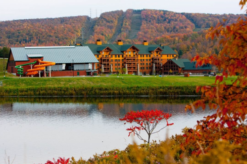 A scenic view of a lodge by a lake, surrounded by autumn foliage and mountains in the background.