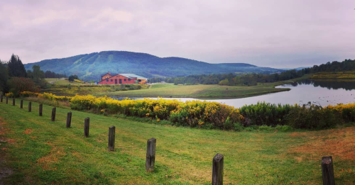 A serene landscape featuring a lake, rolling hills, and a rustic building surrounded by greenery under a cloudy sky.