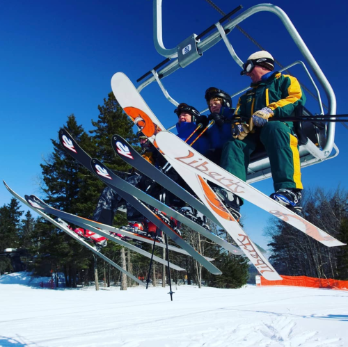 Three skiers on a chairlift, surrounded by snow-covered trees and a clear blue sky.